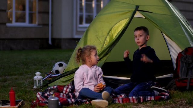 Full shot of young Caucasian brother and sister sitting on blanket in front of camping tent pitched on lawn near suburban family home, girl eating hotdog while boy is fooling around