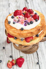 Victoria Sponge Cake with whipped cream, sugared flowers, strawberries and blueberries over a white wooden rustic table top. Image shot for above with blurred background..