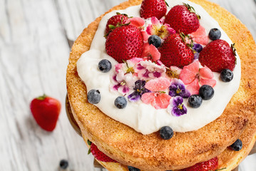 Victoria Sponge Cake with whipped cream, sugared flowers, strawberries and blueberries over a white wooden rustic table top. Image shot for above with blurred background..