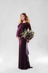 Portrait of young pregnant woman with big flower in her hand. Beautiful model posing in dark dress and holding her stomach at white studio background. Beautiful and happy pregnancy concept