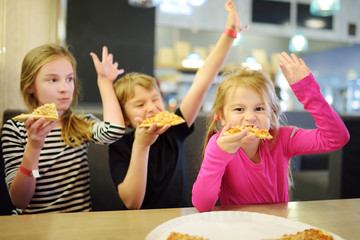 Three cute funny siblings eating slices of pizza at indoor restaurant or cafe.