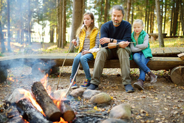 Cute little sisters and their father roasting marshmallows on sticks at bonfire. Children having fun at camp fire. Camping with kids in fall forest.