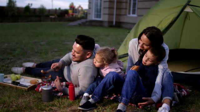 Full shot of loving Caucasian husband and wife with kids sitting on lawn in back yard of suburban home and enjoying evening barbeque, with father roasting sausages on gas cooker while others watch