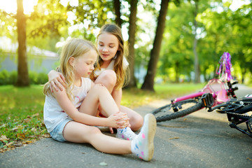 Adorable girl comforting her little sister after she fell off her bike at summer park. Child...