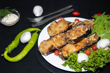 Eggplant stuffed with mushrooms and onions, rubbed with cheese, baked in the oven next to cherry tomatoes, lettuce. In the bowl sauce. All on a black background.