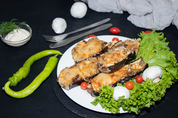 Eggplant stuffed with mushrooms and onions, rubbed with cheese, baked in the oven next to cherry tomatoes, lettuce. In the bowl sauce. All on a black background.