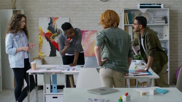 Panning shot of four multiracial creative startupers in their twenties collaborating at loft studio and conceptualizing new project, standing around cluttered table, engaged in animated discussion
