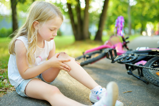 Cute little girl sitting on the ground after falling off her bike at summer park. Child getting hurt while riding a bicycle.