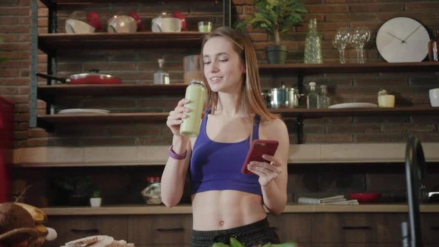 Cheerful young stylish woman in sportswear standing in modern kitchen and actively surfing the internet via phone, then takes a sip of organic smoothie and looks straight to camera, smiling cutely