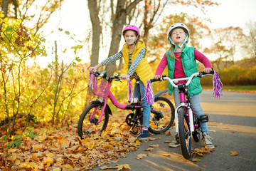 Fototapeta na wymiar Cute little sisters riding bikes in a city park on sunny autumn day. Active family leisure with kids.