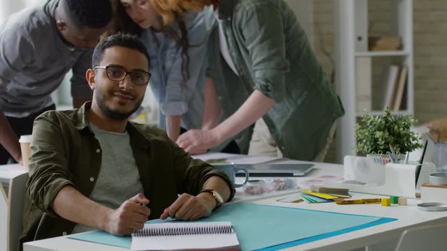 Medium portrait shot of young handsome Middle Eastern illustrator drawing sketch in notebook and smiling warmly for camera, while his colleagues are discussing project materials in background