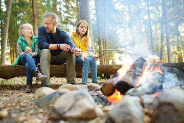 Cute little sisters and their father roasting marshmallows on sticks at bonfire. Children having fun at camp fire. Camping with kids in fall forest.