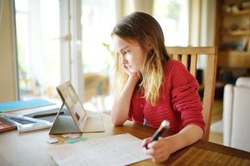 Smart preteen schoolgirl doing her homework with digital tablet at home. Child using gadgets to study.