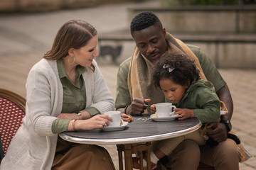 small girl looking at coffee cup sitting on fathers laps in outdoor cafe with mother