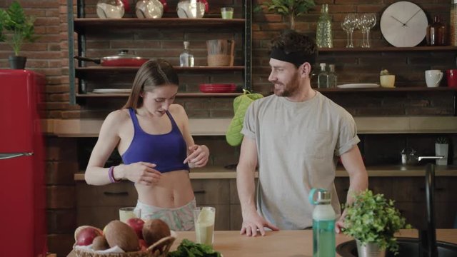 Slow motion of cute young Caucasian couple being in modern kitchen, fit gorgeous woman points on her abs, man takes off his shirt and moves his muscles, both are laughing. Sport, workout concept