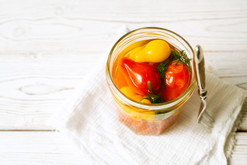 Marinated tomatoes in open glass jar, fork on white wooden background. Horizontal image with copy space