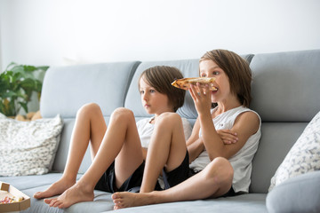 Obraz na płótnie Canvas Cute children, sitting on couch, eating pizza and watching TV. Hungry child taking a bite from pizza on a pizza party