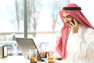 Happy arab man using a laptop and talking on phone in a bar