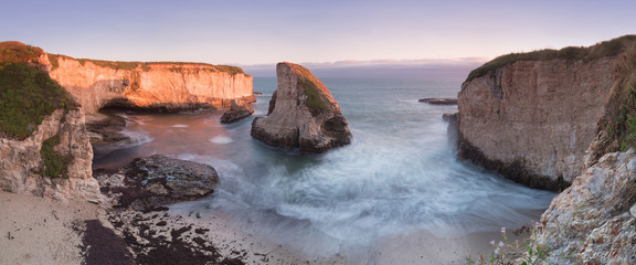 Panoramic view over Shark Fin Cove (Shark Tooth Beach). Davenport, Santa Cruz County, California, USA. Sunset in California - waves and sun hitting these beautiful rock formations with flowers.