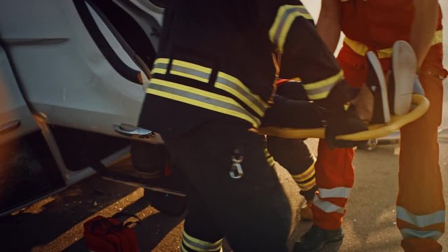 On the Car Crash Traffic Accident Scene: Rescue Team of Firefighters Pull Female Victim out of Rollover Vehicle, They Use Stretchers Carefully, Hand Her Over to Paramedics who Perform First Aid