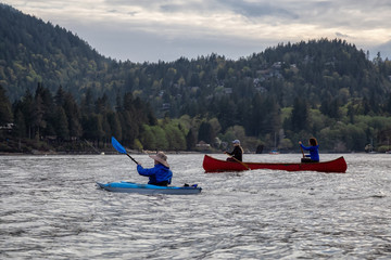 Adventurous friends on a red canoe and kayak are paddling in the Howe Sound during a cloudy sunset....