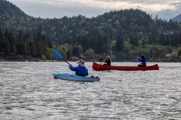 Adventurous friends on a red canoe and kayak are paddling in the Howe Sound during a cloudy sunset....