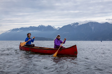 Couple adventurous female friends on a red canoe are paddling in the Howe Sound during a cloudy sunset. Taken near Bowen Island, West of Vancouver, BC, Canada.