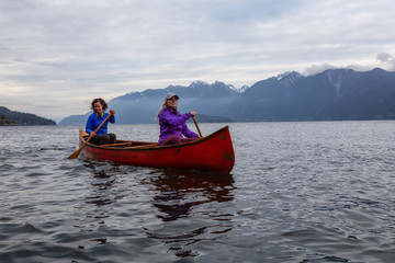 Fototapeta na wymiar Couple adventurous female friends on a red canoe are paddling in the Howe Sound during a cloudy sunset. Taken near Bowen Island, West of Vancouver, BC, Canada.