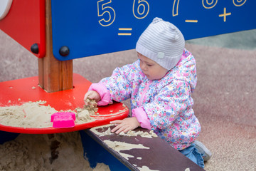Baby girl playing with sand. A girl in a pink jacket and hat for a walk plays with sand in a sandbox, picks up sand, studies the world around.