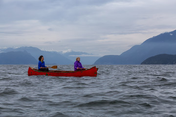 Couple adventurous female friends on a red canoe are paddling in the Howe Sound during a cloudy...