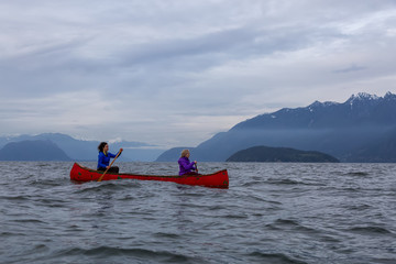 Couple adventurous female friends on a red canoe are paddling in the Howe Sound during a cloudy...