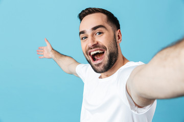 Excited young happy bearded man posing isolated over blue wall background take a selfie by camera.