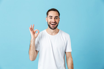 Fototapeta premium Smiling young happy bearded man posing isolated over blue wall background showing okay gesture. Summer concept.