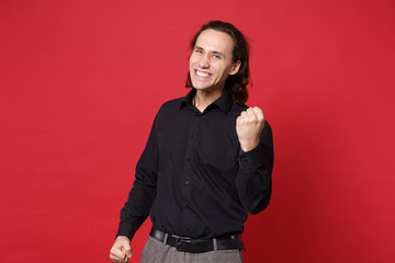 Handsome stylish young curly long haired man in black shirt posing isolated on red wall background studio portrait. People sincere emotions lifestyle concept. Mock up copy space. Doing winner gesture.