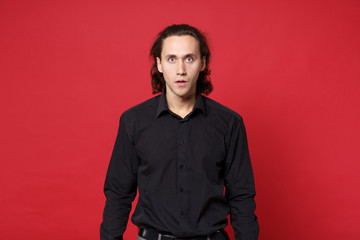 Obraz premium Shocked upset stylish young curly long haired man in black shirt posing isolated on red wall background studio portrait. People sincere emotions lifestyle concept. Mock up copy space. Looking camera.