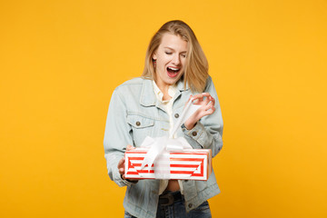 Cheerful young woman in denim casual clothes hold red striped present box with gift ribbon isolated...