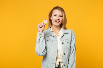 Portrait of stunning young woman in denim casual clothes looking camera holding bitcoin, future currency isolated on yellow orange background in studio. People lifestyle concept. Mock up copy space.