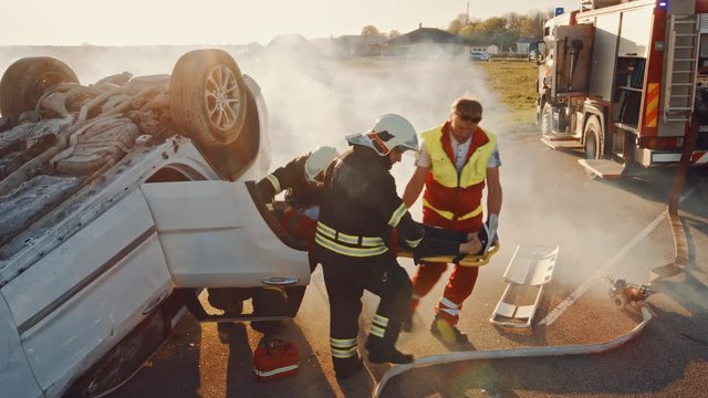 On the Car Crash Traffic Accident Scene: Rescue Team of Firefighters Pull Female Victim out of Rollover Vehicle, They Use Stretchers, Hand Her Over to Paramedics who Do First Aid. High Angle