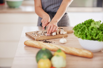 Dedicated Caucasian housewife in apron standing in kitchen and chopping mushrooms. On table are lots of vegetables. Cooking at home concept.