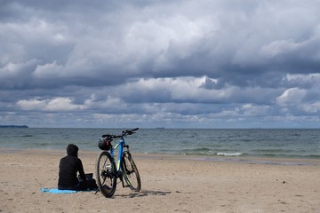 Fototapeta premium Poland, Gdansk, Baltic Sea - women in black cloth sitting on the beach with bike in cloudy day with amazing dramatic clouds