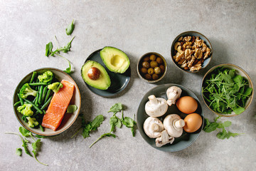 Ketogenic diet ingredients for cooking dinner. Raw salmon, avocado, broccoli, bean, olives, nuts mushrooms, eggs in ceramic bowls. Grey texture background. Flat lay, space