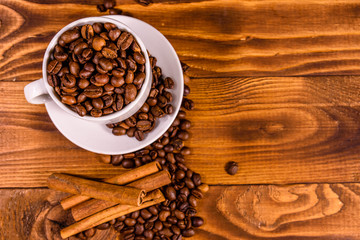 White cup filled with coffee beans and cinnamon sticks on wooden table. Top view