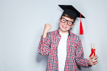Young boy in graduation cap with diploma on grey background
