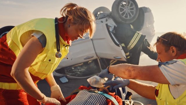 On the Car Crash Traffic Accident Scene: Paramedics Save Life of a Female Victim Lying on Stretchers. They Listen To a Heartbeat, Apply Oxygen Mask and Give First Aid. Firefighters Extinguish Fire