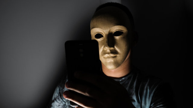 Male hacker hidden face with the mask accessing to personal information on laptop (phone) in the dark. Technology, cyber crime concept. 