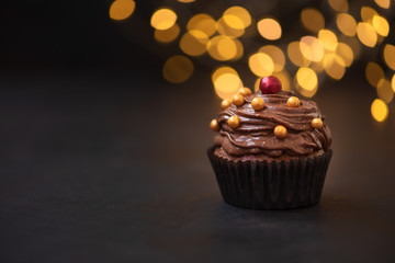 Chocolate cupcake with golden candies on dark wooden background against blurred lights. Selective focus. Unhealthy food.