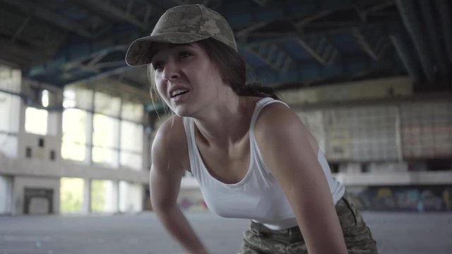 Confident young woman in military uniform training in dusty dirty abandoned building. Slim girl running up to the camera and stopping to catch her breath. Warrior woman in a deserted factory.