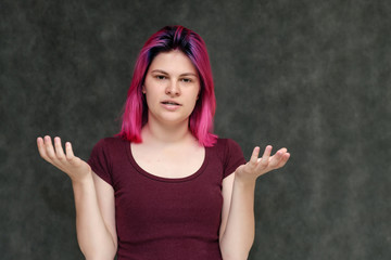 Portrait to the waist of a young pretty girl teenager in a burgundy T-shirt with beautiful purple hair on a gray background in the studio. Talking, smiling, showing hands with emotions.