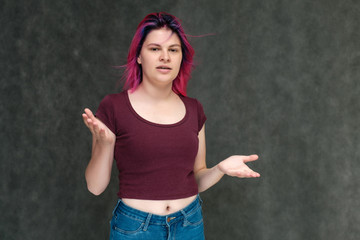 Portrait to the waist of a young pretty teen girl in a burgundy T-shirt and jeans with beautiful purple hair on a gray background in the studio. Talking, smiling, showing hands with emotions.