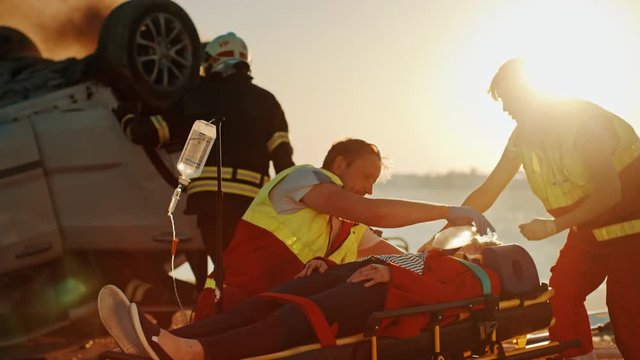 On the Car Crash Traffic Accident Scene: Paramedics Save Life of a Passenger Lying on Stretchers. They Listen To a Heartbeat, Use Oxygen Mask, setup Drop Counter. Firefighters Use Cutters to Open Car